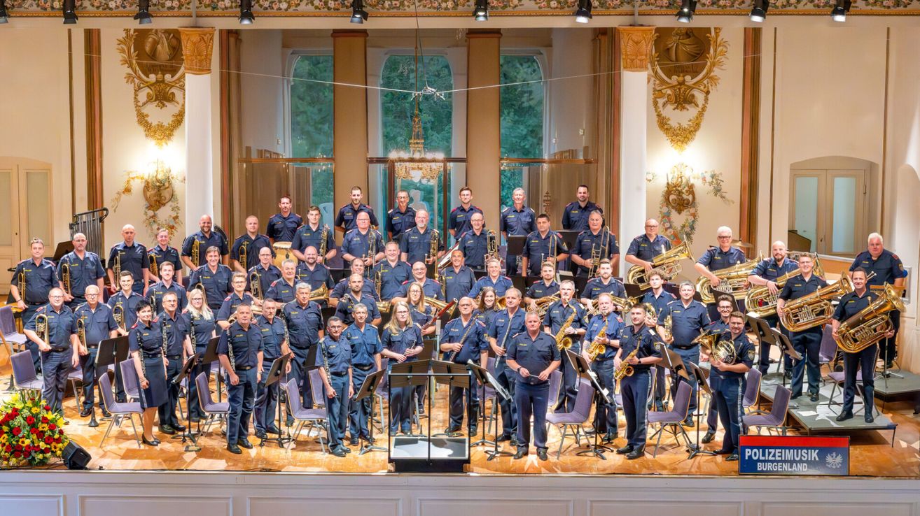 Eine große Gruppe uniformierter Personen, möglicherweise eine Band, steht auf einer Bühne mit Musikinstrumenten. Die Bühne ist mit aufwendigen Säulen und einer Kristallleuchter dekoriert.