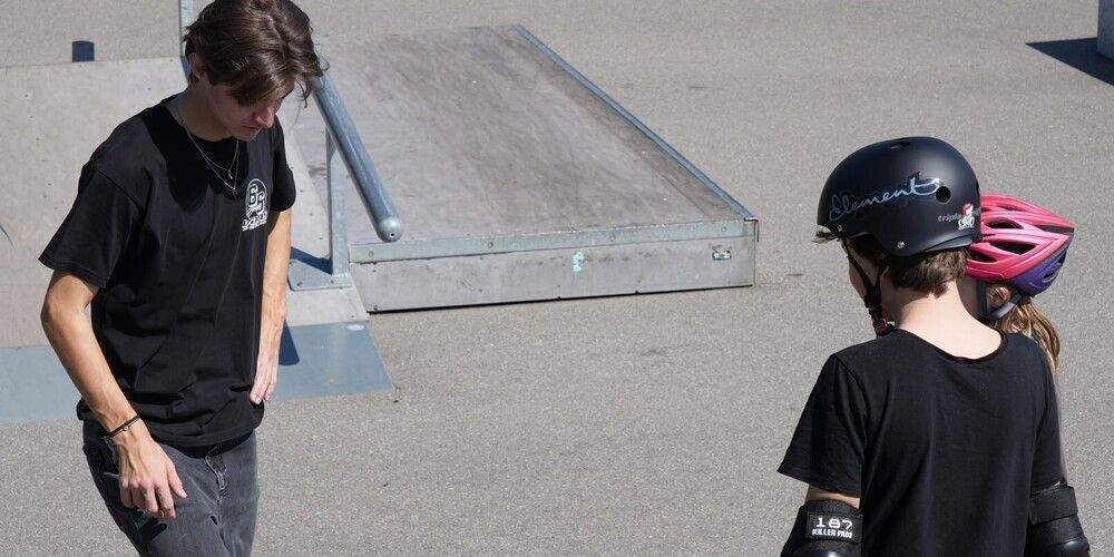 Two young men stand at a skate park, one wearing a helmet and elbow pads, the other with a necklace. Behind them is a ramp.