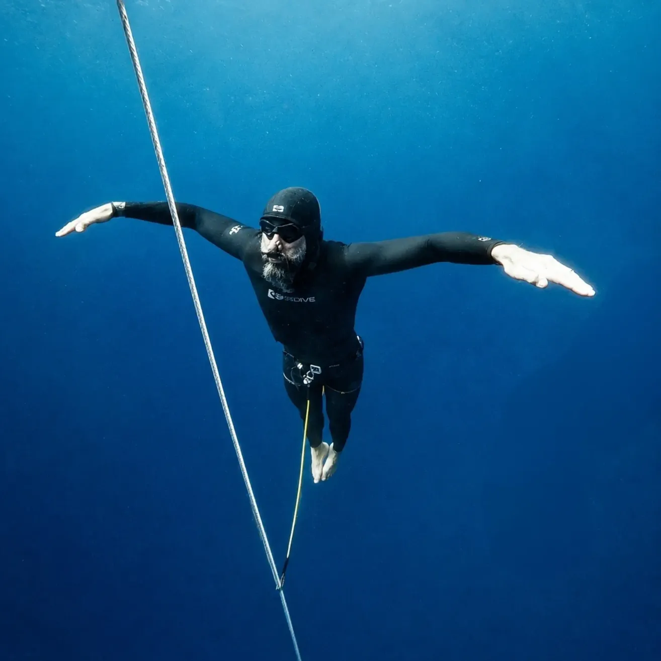 Ein Mann in einem schwarzen Neoprenanzug und Taucherbrille hängt unter Wasser und hält sich an einem Seil fest. Er scheint ein professioneller Taucher zu sein.