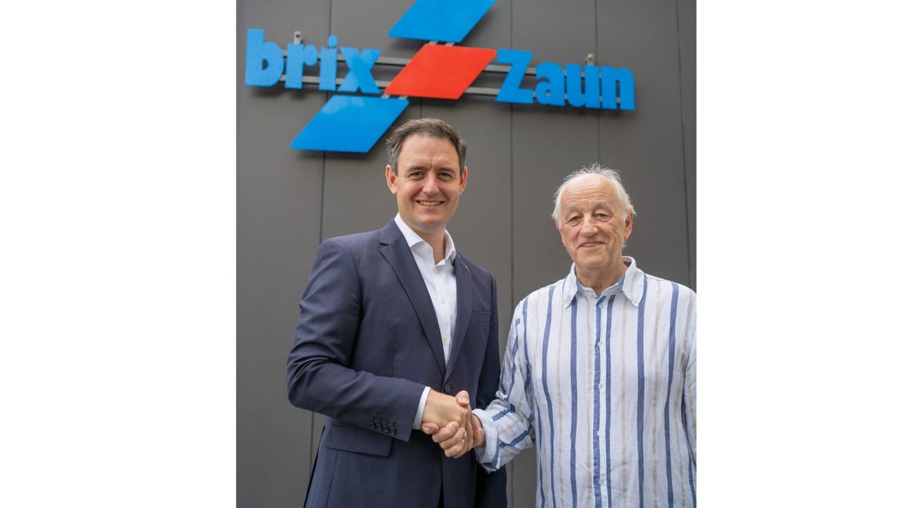 Two men are standing in front of a Brix Zaun sign, smiling and shaking hands.