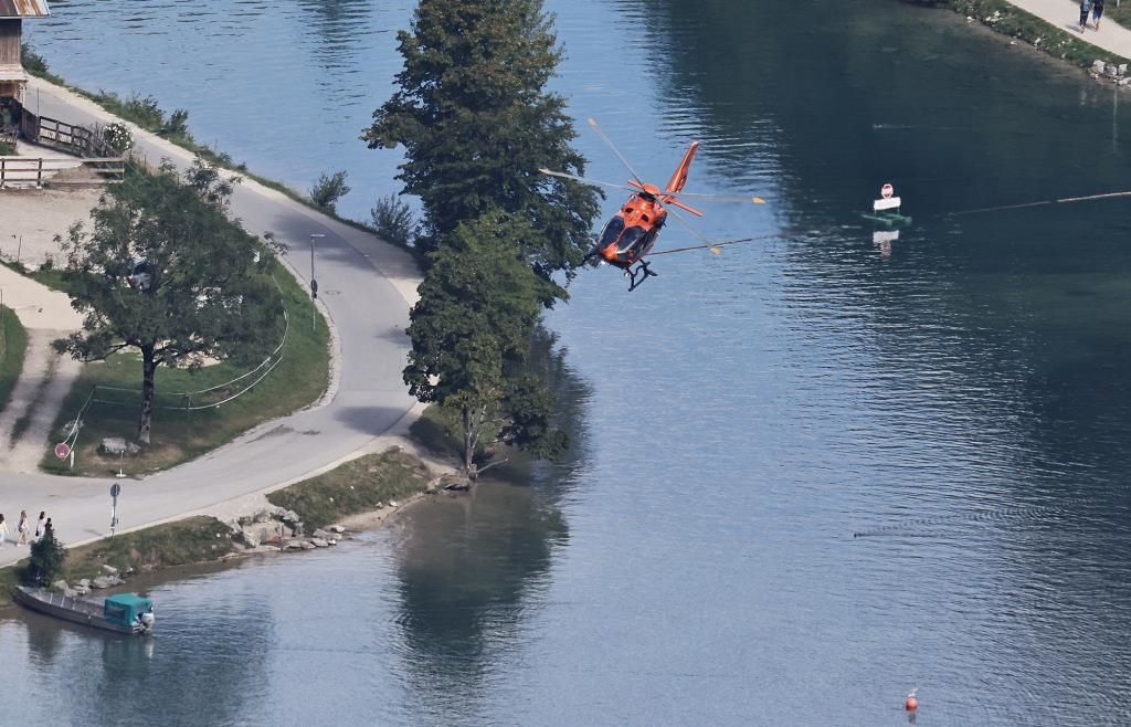 Ein roter Hubschrauber schwebt über einem See, möglicherweise im Einsatz für eine Rettungsaktion. Bäume und eine Straße sind links zu sehen.