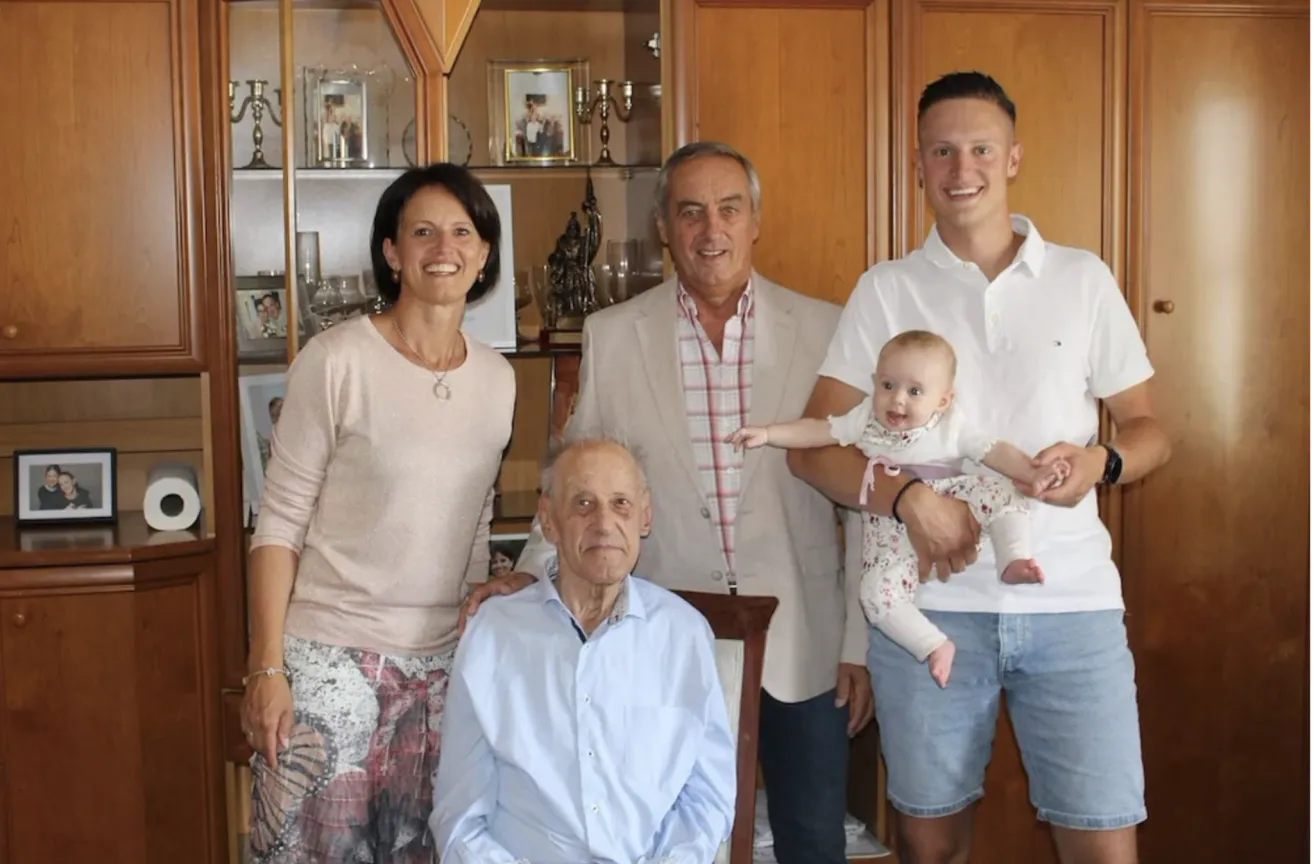 Ein Familienfoto mit einer lächelnden Frau, einem älteren Mann und zwei Männern, die ein Baby in einem Wohnzimmer mit Holzschränken und Bilderrahmen halten.
