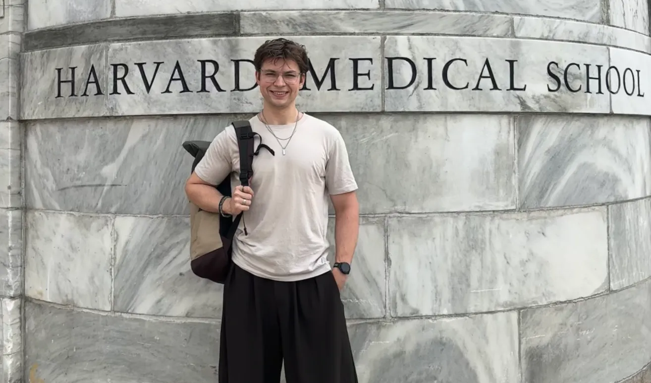 A man with glasses and a necklace stands in front of a marble wall with the word 'Medical' written on it. He is wearing a white t-shirt, black pants, and a backpack.