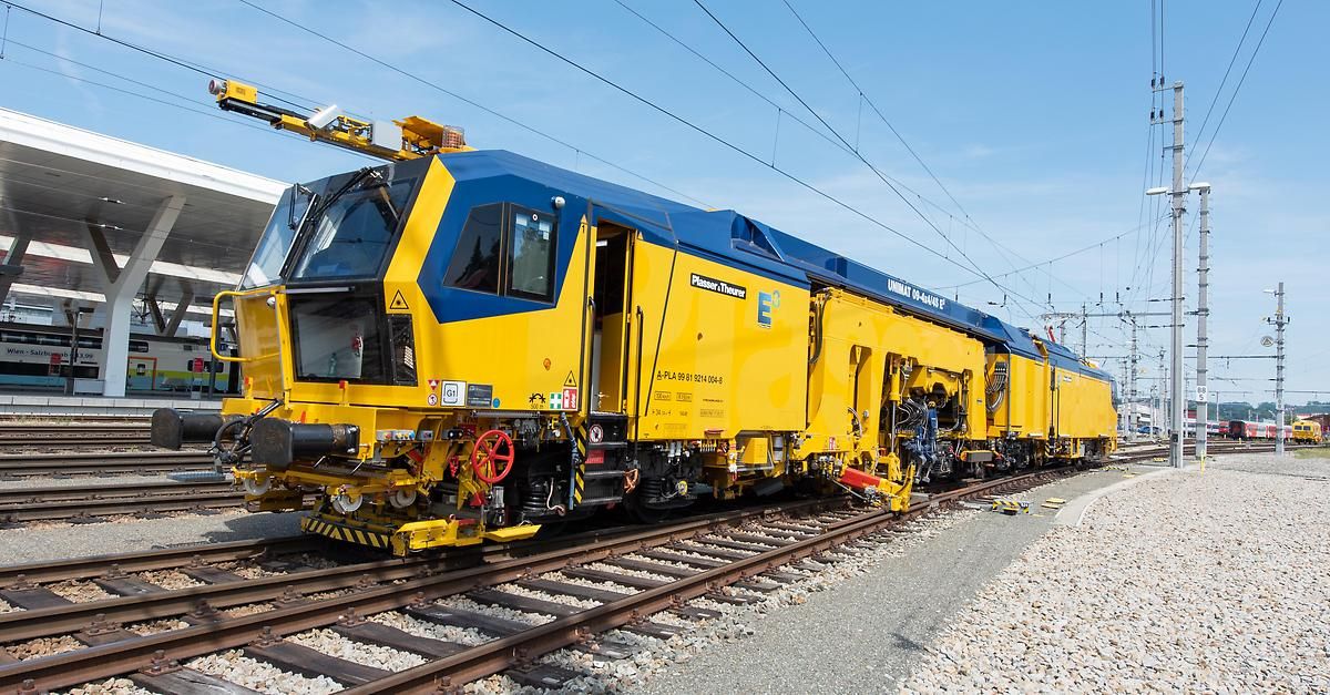 A yellow and blue train with a large red wheel on the front is parked on the railway track. The train has a large open door and a yellow crane on top.