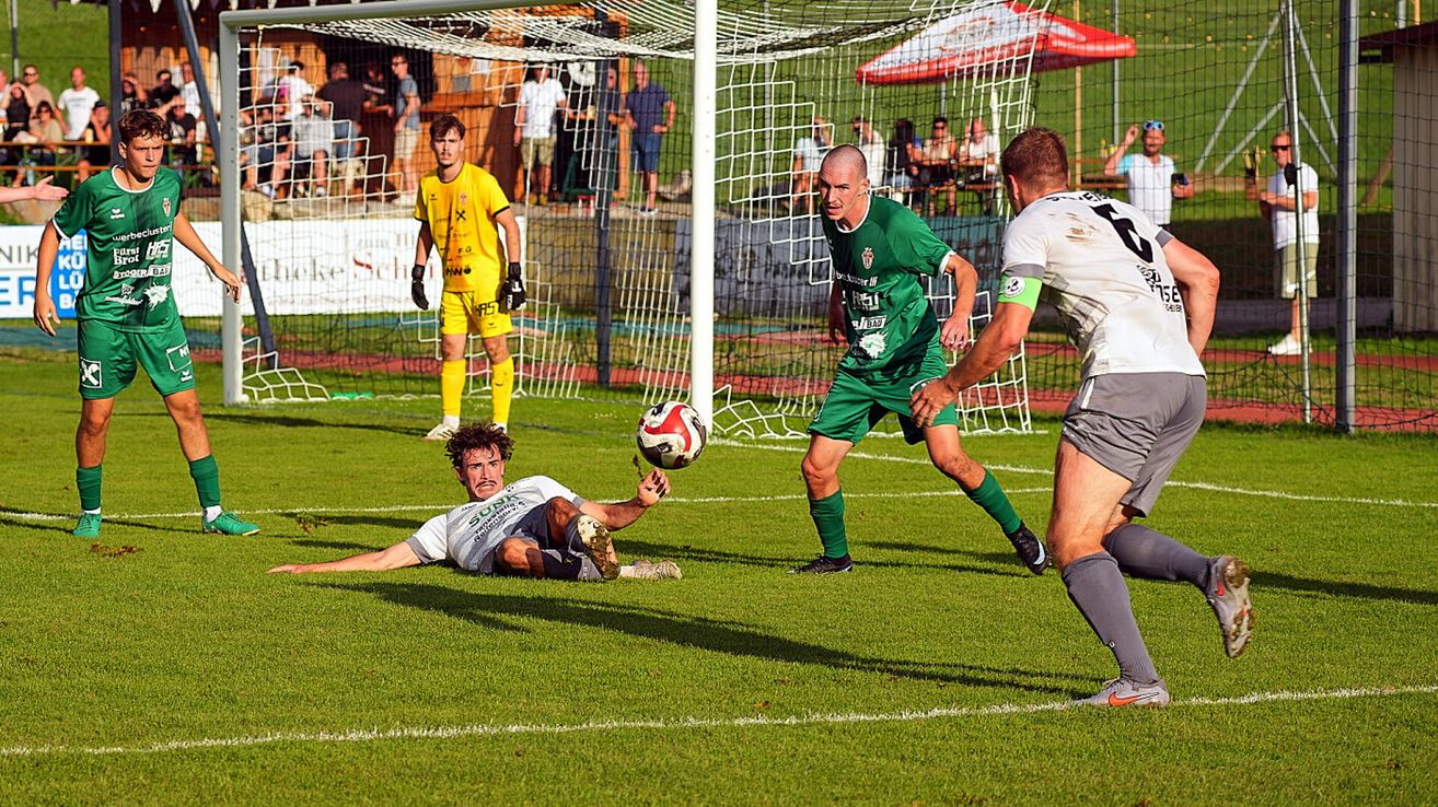 Ein Fußballspieler in einem weißen Trikot taucht ab, um den Ball zu blockieren, während ein anderer Spieler in Grün versucht, ihn zu schießen. Ein Torwart in Gelb steht hinter dem Netz.