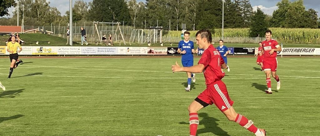 Ein Fußballspiel findet auf einem Feld statt, auf dem Spieler in roten und blauen Uniformen spielen. Der Spieler in Rot läuft auf den Ball zu, während der Spieler in Blau ihm folgt. Es gibt Menschen, die am Rande des Feldes stehen und sitzen.