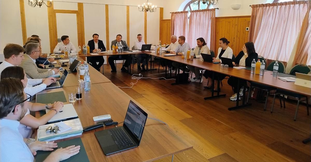 Eine Gruppe von Menschen sitzt um einen langen Tisch in einem Besprechungsraum, mit Laptops, Wasserflaschen und Papieren vor ihnen. Der Raum hat Holzwände und -böden, und es hängen Kronleuchter von der Decke.