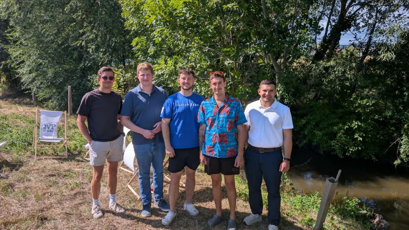Fünf Männer stehen zusammen in einer sonnigen Außenumgebung und lächeln für ein Foto. Sie sind lässig gekleidet und stehen vor einem kleinen Bach.