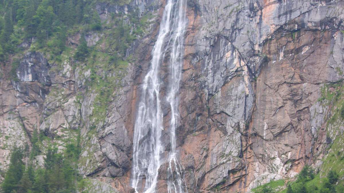 Ein Wasserfall stürzt an einem felsigen Abhang hinunter, umgeben von üppigem grünem Bewuchs und einer bergigen Kulisse.