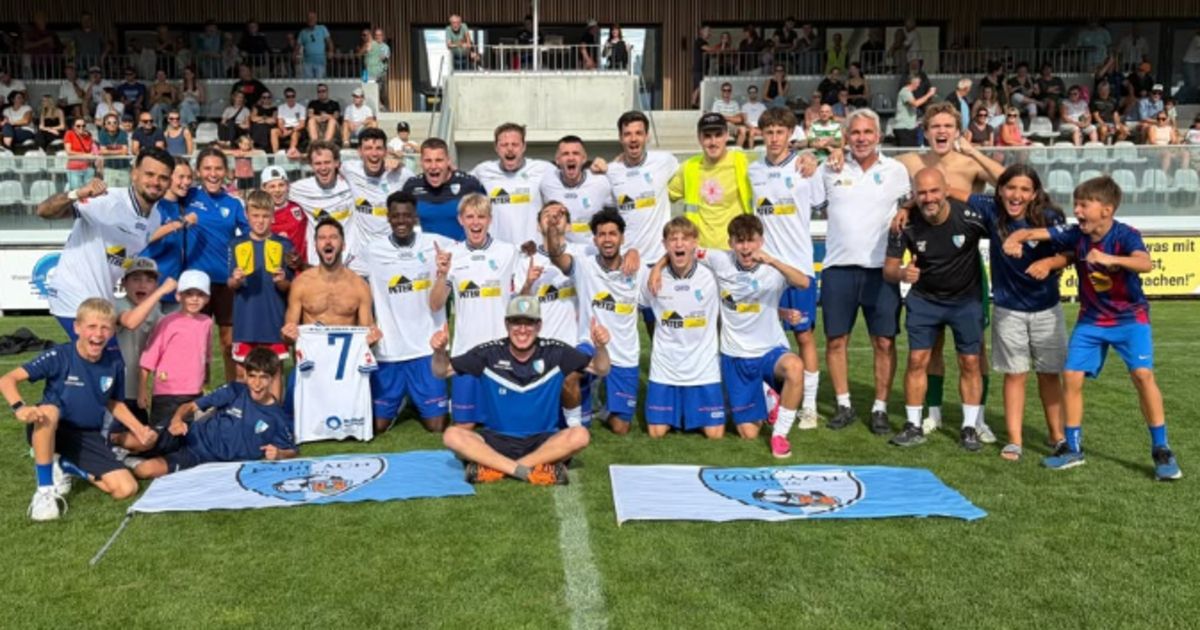 A soccer team poses for a group photo on the field with a cheering crowd in the background.