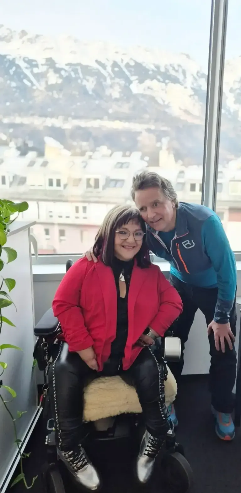 Ein Mann und eine Frau lächeln und posieren für ein Foto in einem Büro mit einem Fenster, das eine Stadtlandschaft im Hintergrund zeigt.