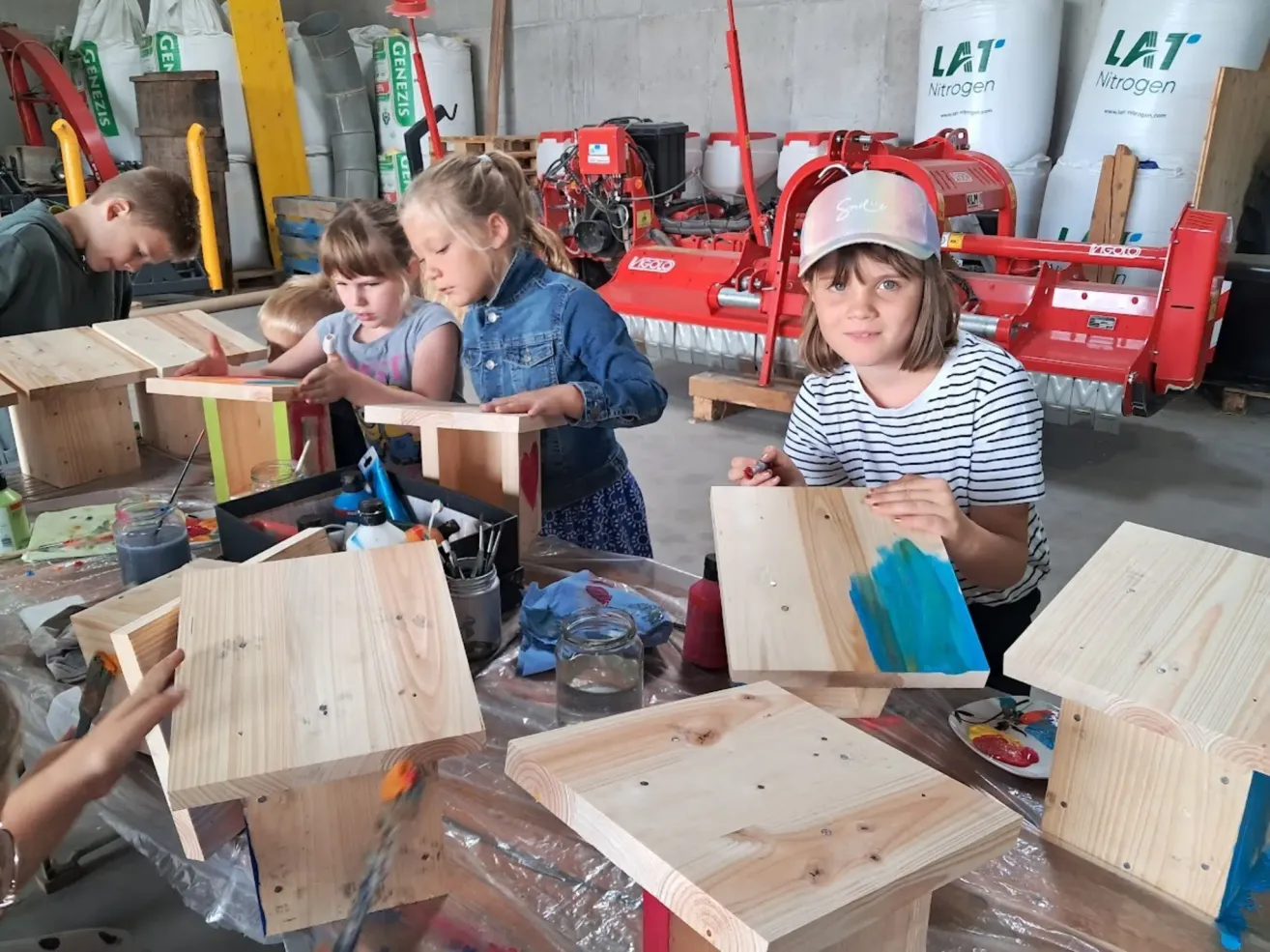 Three young girls are painting wooden birdhouses in a workshop. Each girl is focused on her own birdhouse, with various paint colors and tools scattered around.