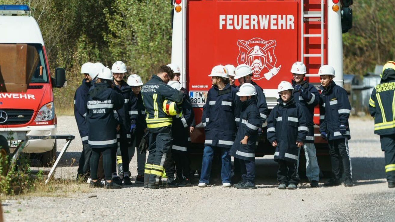 A group of firefighters and children in uniforms standing in front of a fire truck. The firefighters are explaining something to the children.