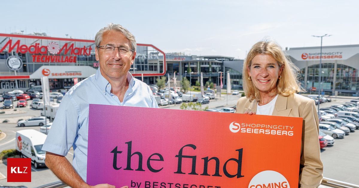 A man and a woman are standing in front of a shopping mall, holding a sign that reads 'the find by BEST SECRET'. The man is wearing glasses and a light blue shirt, while the woman is wearing a beige blazer. Behind them is a parking lot with several cars and a building.
