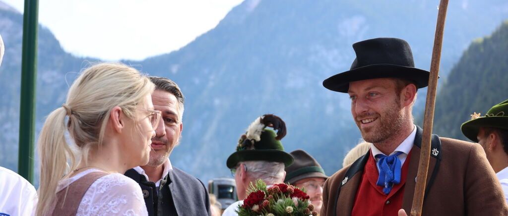 Ein lächelnder Mann in traditioneller Kleidung hält einen Blumenstrauß, neben einer Frau in einem weißen Kleid, mit einem Mann in Anzug dahinter, und einer Bergkulisse im Hintergrund.