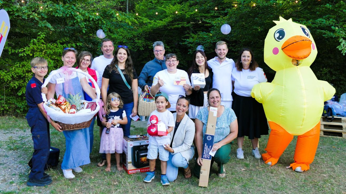 Eine Gruppe von Erwachsenen und Kindern posiert für ein Foto in einem Garten mit einem Korb voller Geschenke und einer Kiste mit Spielzeug im Vordergrund.