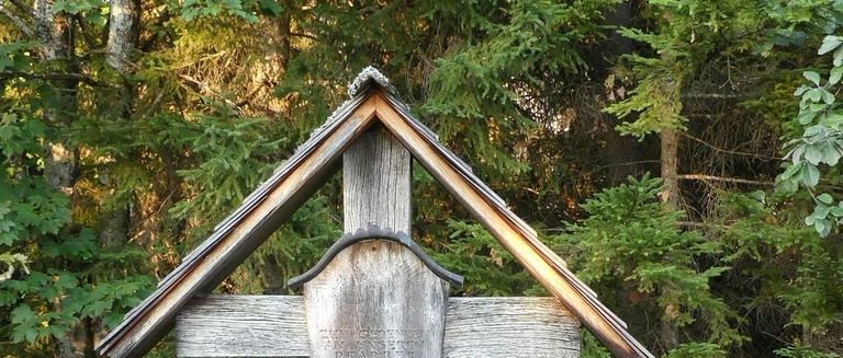 Ein hölzernes Kreuz mit einem geneigten Dach steht in einem Wald, umgeben von grünen Bäumen. Das Kreuz hat eine Metallhalterung am Boden und eine Inschrift auf der Vorderseite.