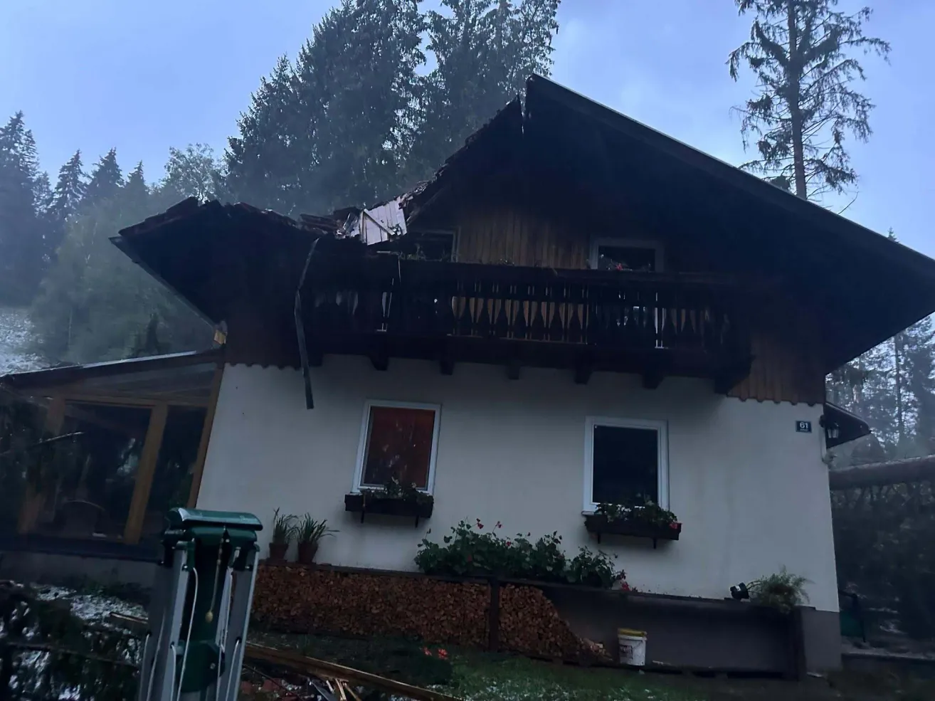 Ein Haus mit einem beschädigten Dach und einem Balkon, umgeben von Bäumen und Pflanzen, mit einem Eimer und einer Leiter in der Nähe.