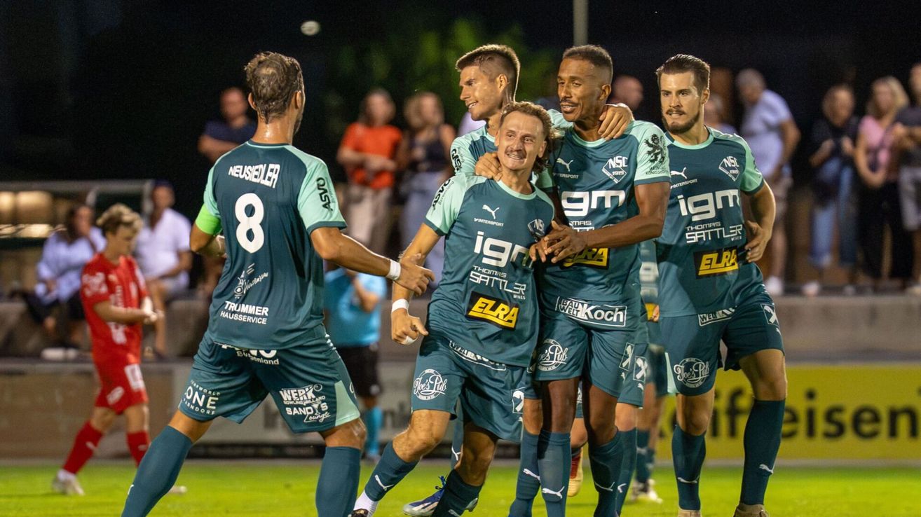 Fünf Fußballspieler in grünen Trikots feiern ein Tor auf dem Feld, wobei einer in der Mitte lächelt und andere um ihn herum sind. Zuschauer sind im Hintergrund zu sehen.