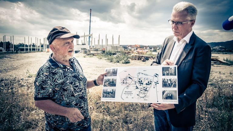 Zwei Männer stehen auf einem Feld, einer hält eine Karte und der andere schaut darauf. Sie stehen vor einer Baustelle mit einem Turm und Maschinen.