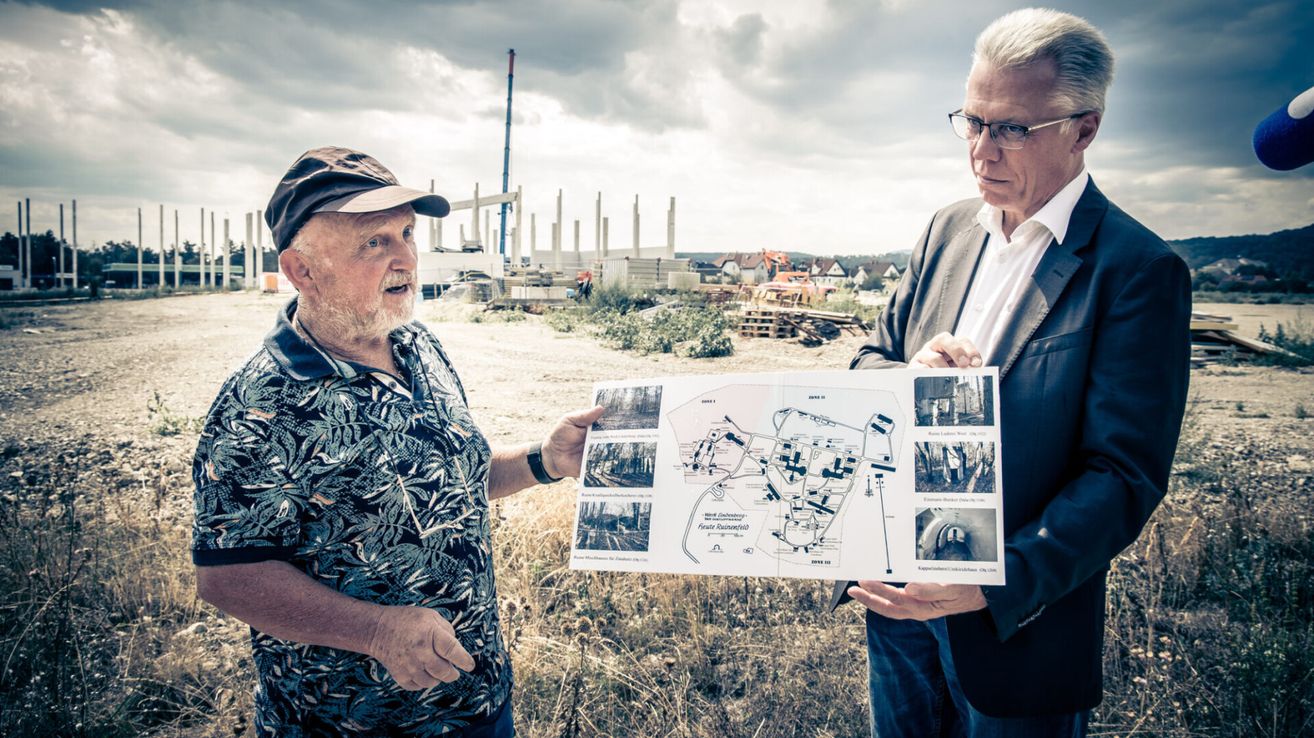 Zwei Männer stehen auf einem Feld, einer hält eine Karte und der andere schaut darauf. Sie stehen vor einer Baustelle mit einem Turm und Maschinen.