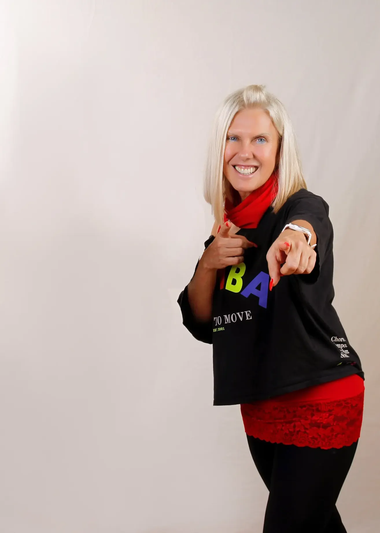 Eine Frau mit blonden Haaren, die ein schwarzes T-Shirt mit bunten Buchstaben und einem roten Schal trägt, posiert lächelnd für ein Foto.
