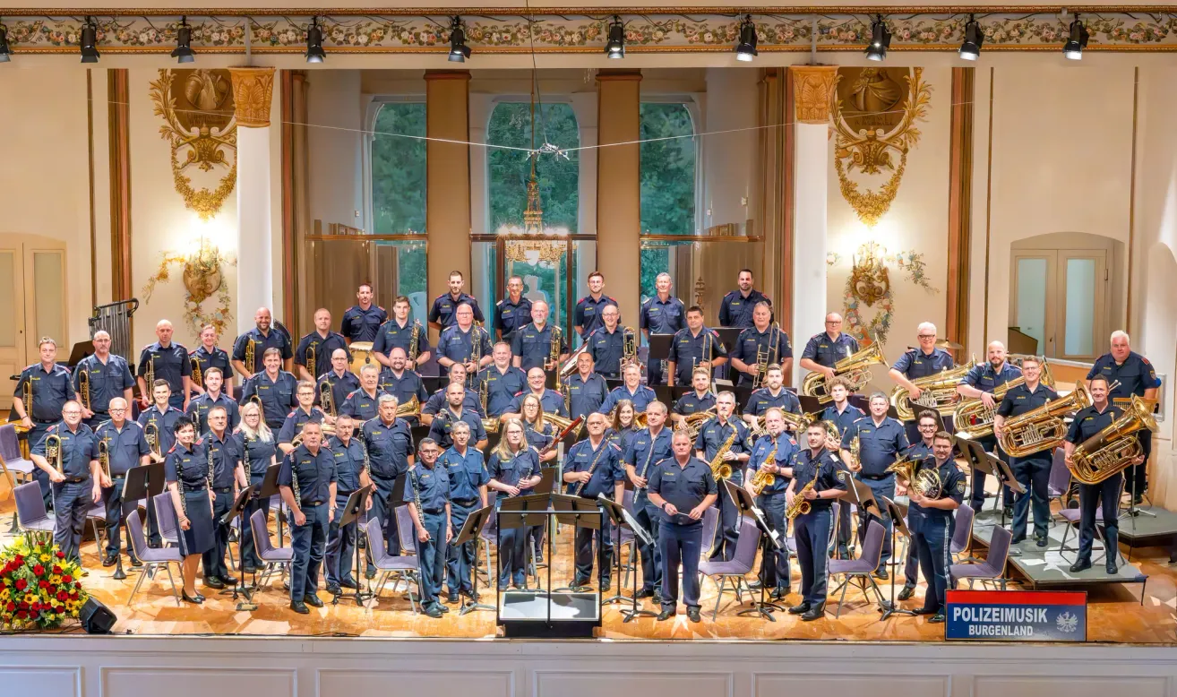 Eine große Gruppe von Musikern in Uniform, die verschiedene Instrumente halten, steht auf einer Bühne mit einem dekorativen Hintergrund und großen Fenstern.