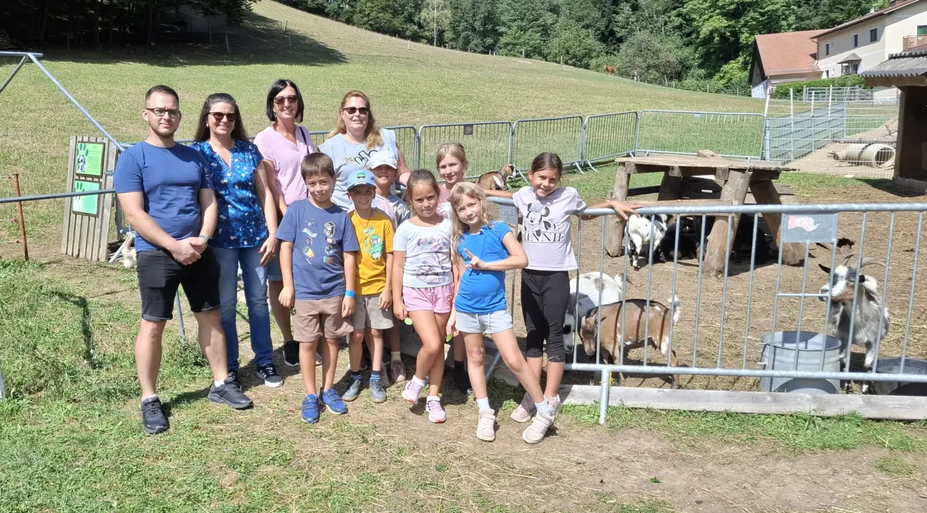 Eine Gruppe von Kindern und Erwachsenen steht vor einem Zaun mit Ziegen auf einem Bauernhof. Die Kinder lächeln und posieren für ein Foto.