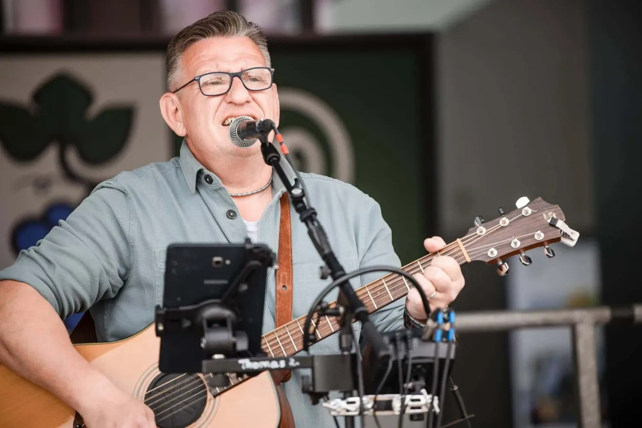 Ein Mann mit Brille und Halskette spielt Gitarre und singt in ein Mikrofon. Er hält die Gitarre mit beiden Händen und hat einen Gurt um die Schulter.