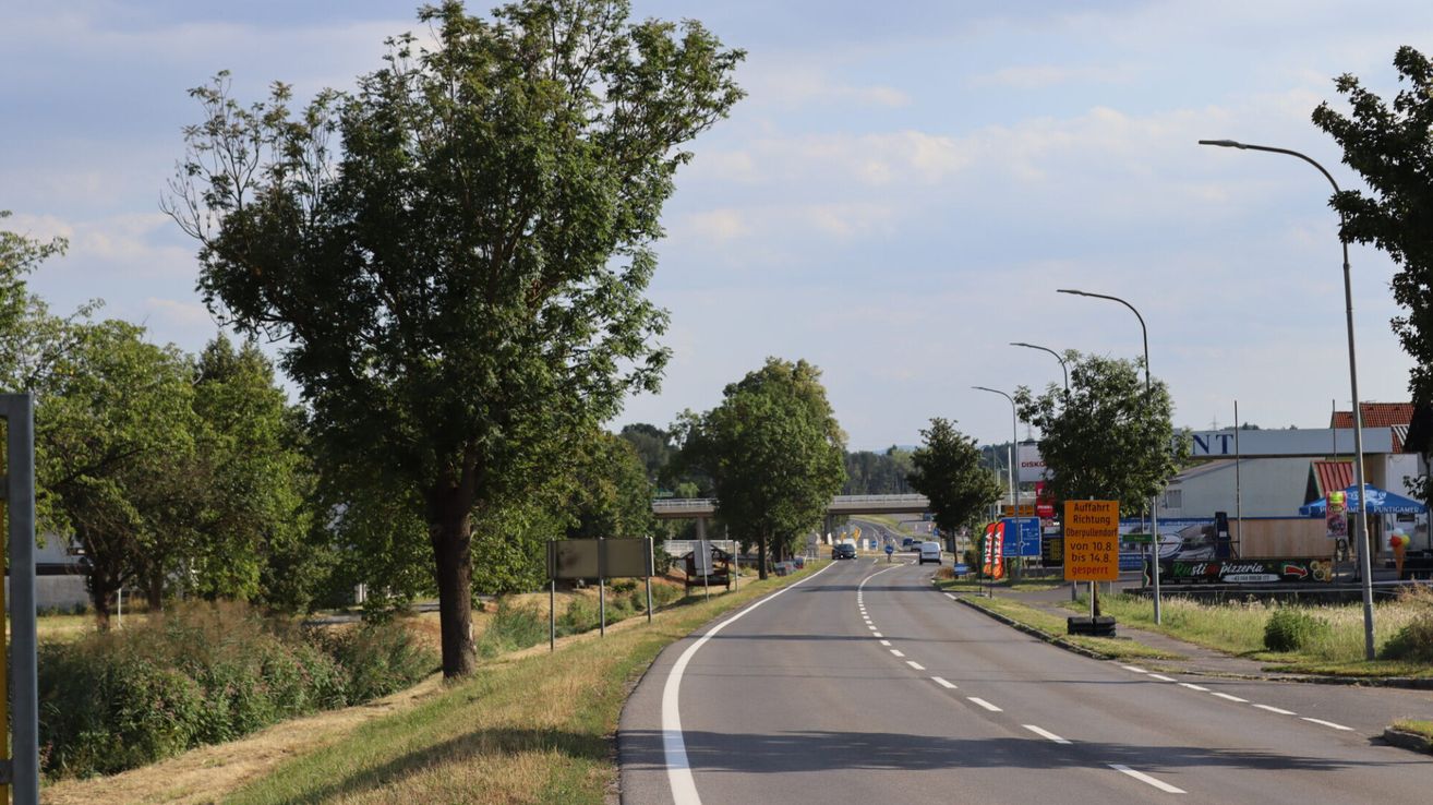 Eine Autobahn mit wenig Verkehr unter einem teilweise bewölkten Himmel, mit Bäumen, Straßenschildern und einer Brücke in der Ferne.
