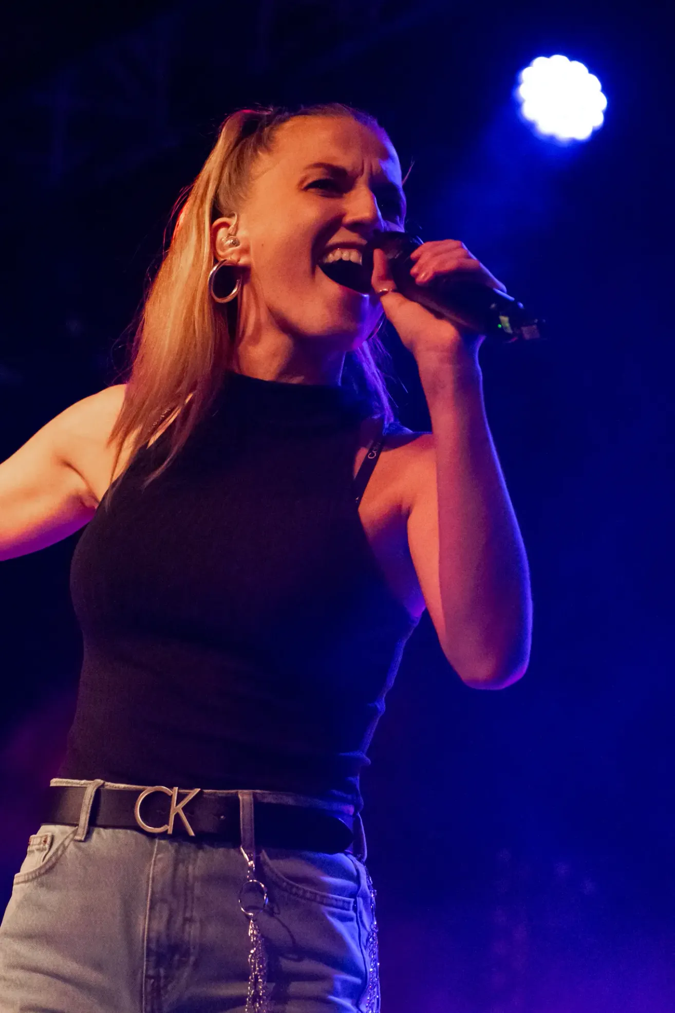 A woman with blonde hair is singing into a microphone on stage. She is wearing a black sleeveless top and a belt with the letters CK.