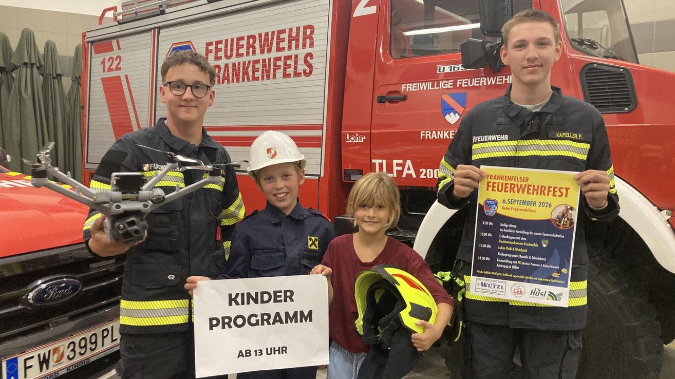 Drei Kinder in Feuerwehruniformen stehen vor einem Feuerwehrauto und halten Schilder. Der Junge auf der linken Seite hält ein Schild mit der Aufschrift 'KINDER PROGRAMM AB 13 UHR'. Der Junge in der Mitte hält einen Helm. Der Junge auf der rechten Seite hält ein Plakat.