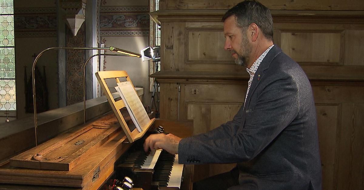 Ein Mann in einem Anzug spielt eine Orgel in einem hölzernen Raum, mit einem Notenständer vor ihm.