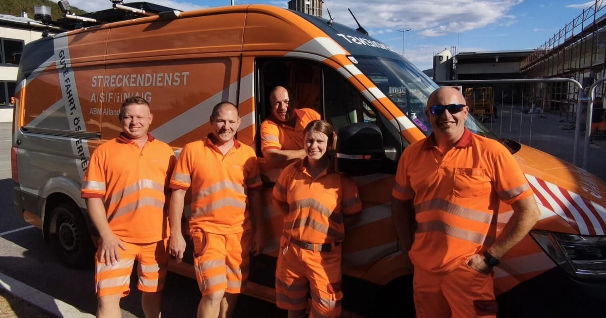 Fünf Personen in orangefarbenen Uniformen stehen vor einem orangefarbenen Van mit dem Wort 'Technische' darauf. Sie lächeln und scheinen für ein Foto zu posieren.