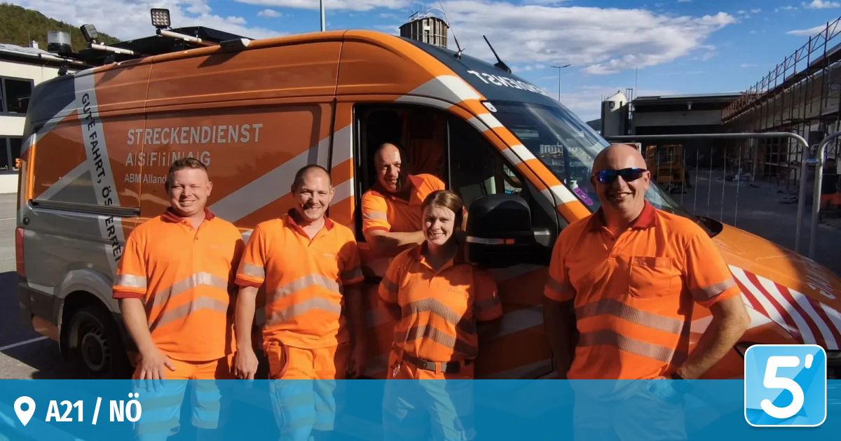 Fünf Personen in orangefarbenen Uniformen stehen vor einem großen orangefarbenen Fahrzeug mit dem Wort 'Transport' darauf. Der Himmel ist klar mit einigen Wolken.