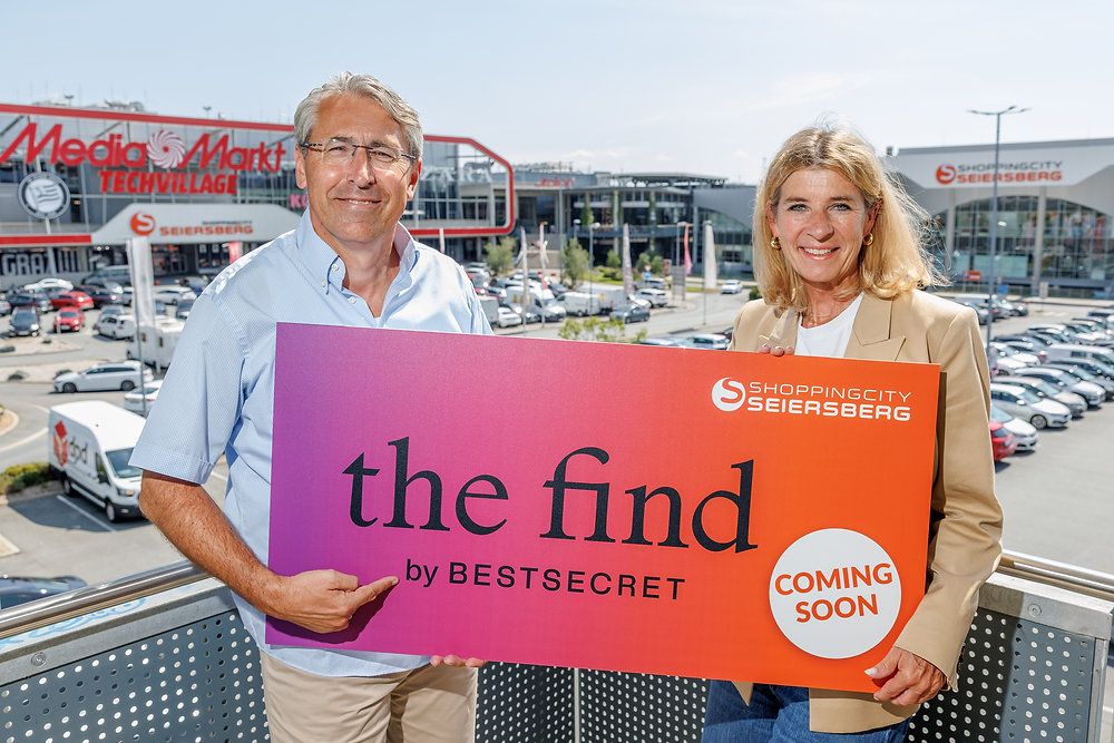 A man and a woman stand on a balcony holding a large sign that reads 'the find by BESTSECRET' with a pink and orange background. The sign also includes a white circle with the text 'COMING SOON'. Behind them, a shopping mall with a large sign reading 'Markt' is visible.
