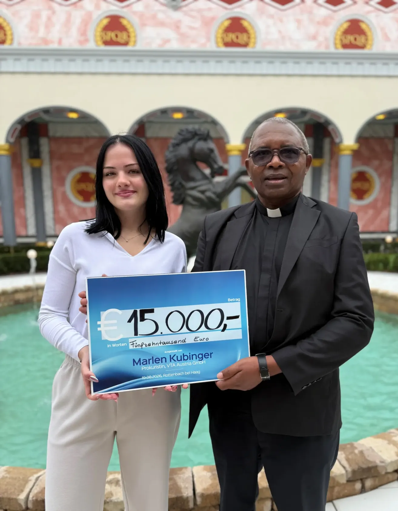 Ein Mann und eine Frau stehen vor einem Brunnen und halten ein blaues Schild mit dem Text '15.000 Euro' und dem Namen 'Marlen Kubinger'. Der Mann trägt einen schwarzen Anzug und eine Brille, und die Frau trägt ein weißes Hemd und Hose. Hinter ihnen steht eine Pferdestatue und ein Gebäude mit Bogenfenstern.