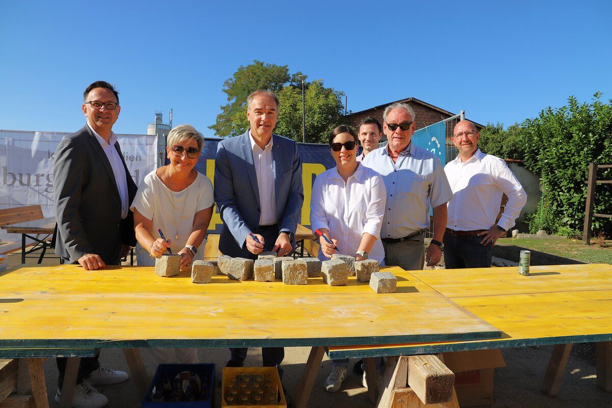 A group of people are standing around a yellow table with concrete blocks, possibly at a construction site. They are signing a document with pens.