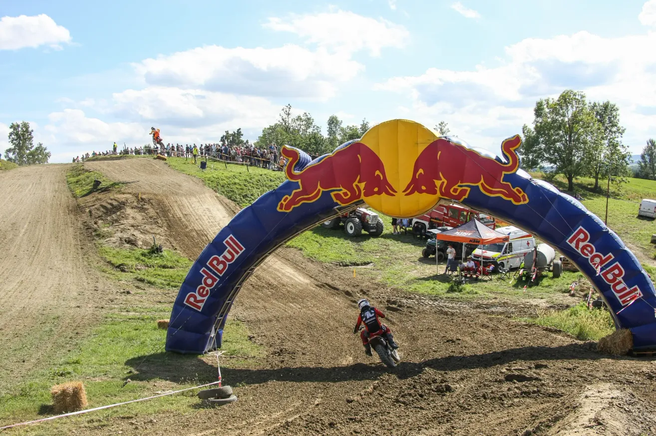 A dirt bike rider wearing a helmet rides through a Red Bull archway on a dirt track with a crowd watching.