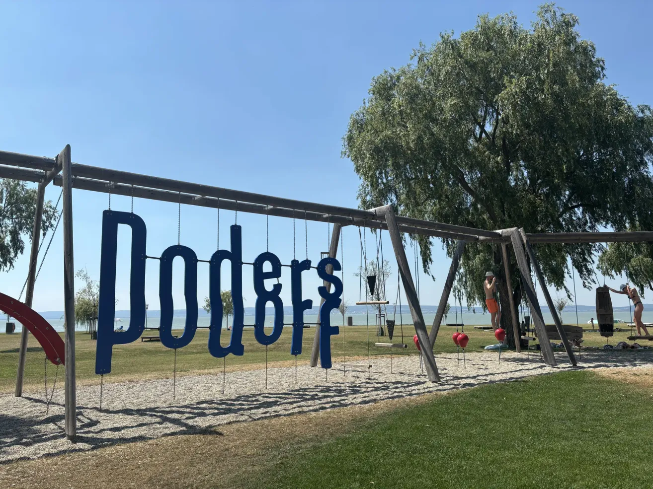 Ein Spielplatz mit Schaukeln und einem Schild mit der Aufschrift 'Poders' unter einem klaren Himmel in der Nähe eines Sees.