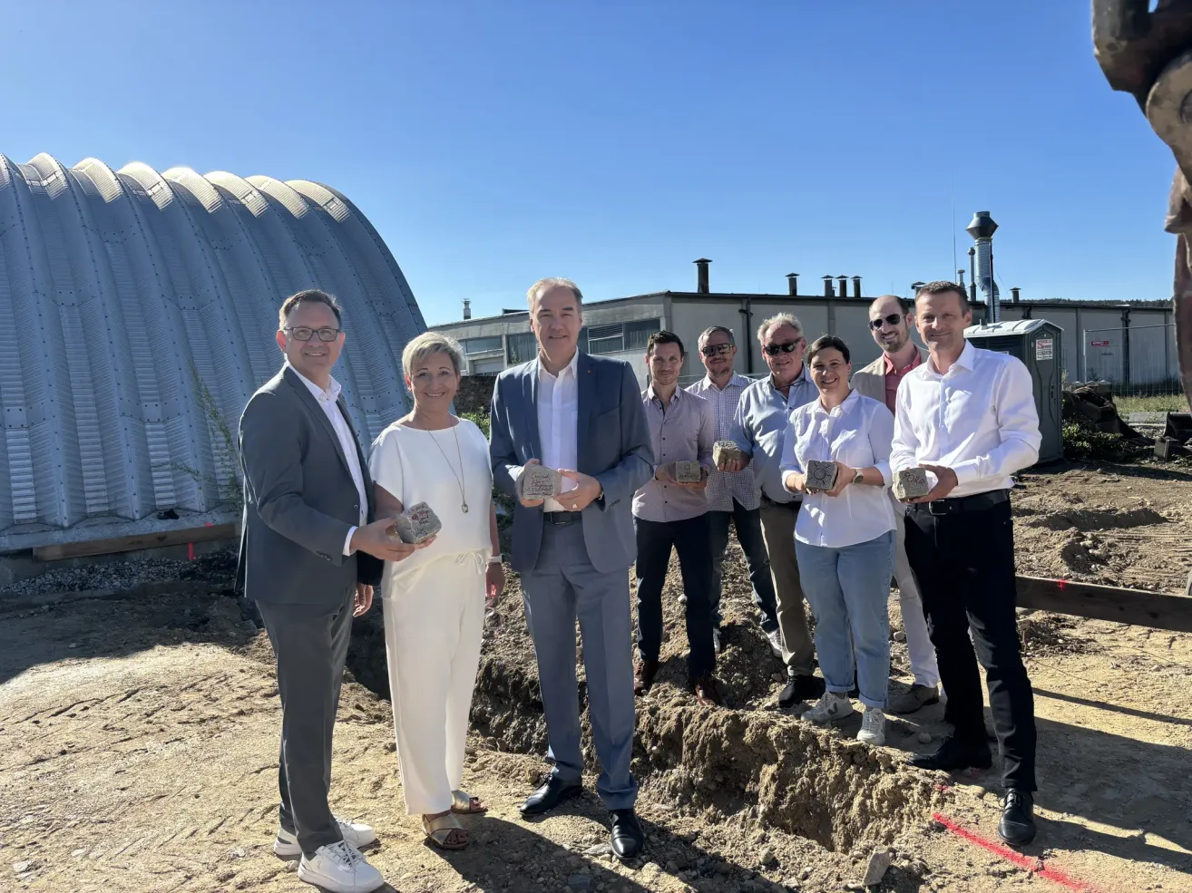 A group of people in formal attire are standing in a dirt area, holding small bricks, and posing for a photo. Behind them, there is a large building with a curved roof.