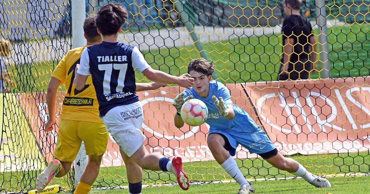 A soccer player in a blue jersey is diving to catch the ball while another player in a yellow jersey tries to block him. The goalkeeper is wearing gloves and sneakers.