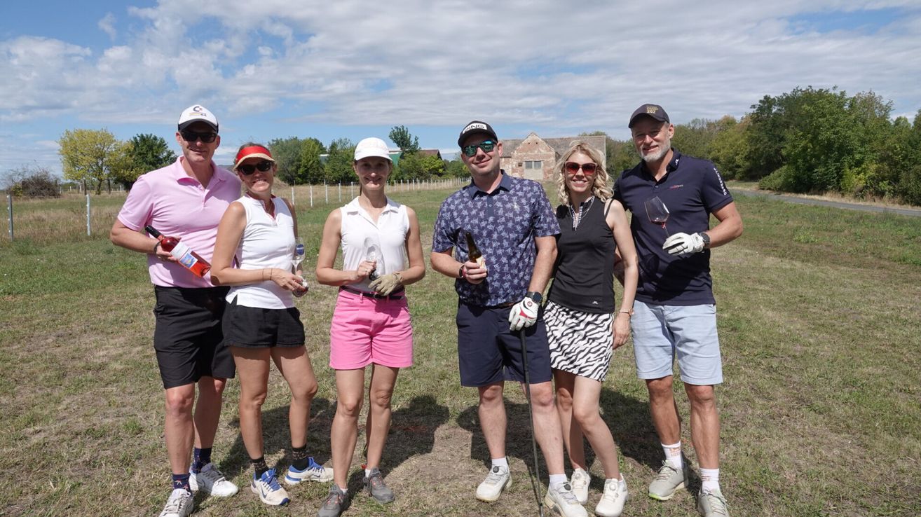 Eine Gruppe von Menschen in Golfkleidung steht auf einem Feld, lächelt und hält Weinglas und Flaschen. Sie posieren für ein Foto mit einem Gebäude im Hintergrund.