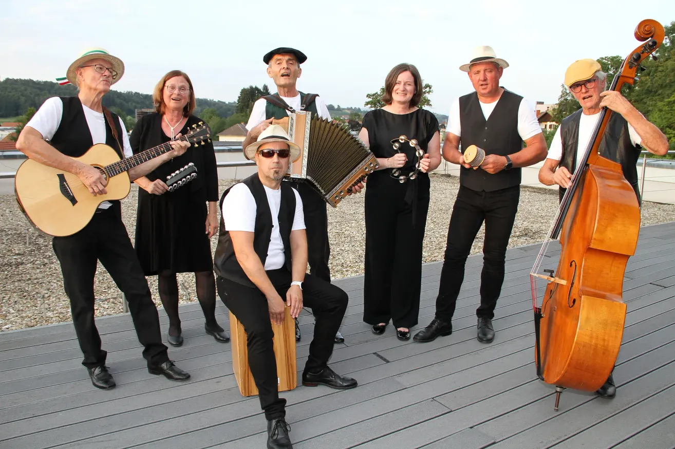 Eine Gruppe von Musikern, in Schwarz gekleidet, posiert für ein Foto auf einer Holzterrasse. Ein Mann sitzt auf einem Holzhocker, während ein anderer ein Akkordeon hält.