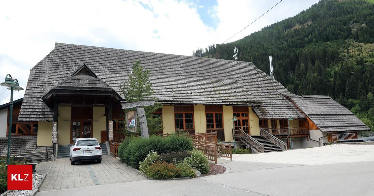 A yellow building with a gray roof and wooden front porch is situated in front of a mountain. A car is parked in front of the building.