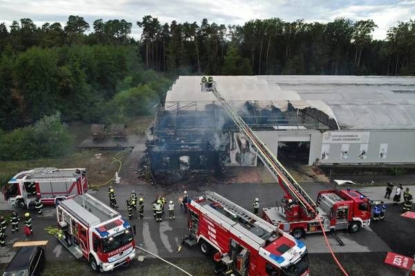 Feuerwehrleute bekämpfen einen Brand in einem Lagerhaus, mit mehreren Feuerwehrwagen und Personal vor Ort. Rauch steigt aus dem Gebäude auf, und eine Leiter ist zur Dachseite hin ausgestreckt.