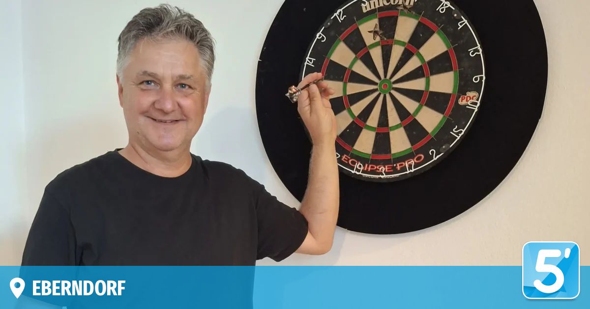 Ein Mann in einem schwarzen T-Shirt hält einen Dart und zielt auf ein Dartboard, das an einer weißen Wand montiert ist.