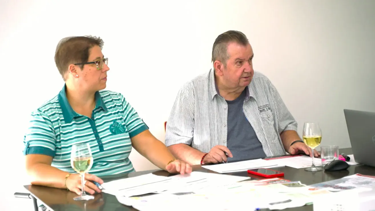 A man and a woman are sitting at a table, facing each other. The man is holding a pen and appears to be writing on a piece of paper. The woman is holding a wine glass and is looking at the man. There are papers and a phone on the table.
