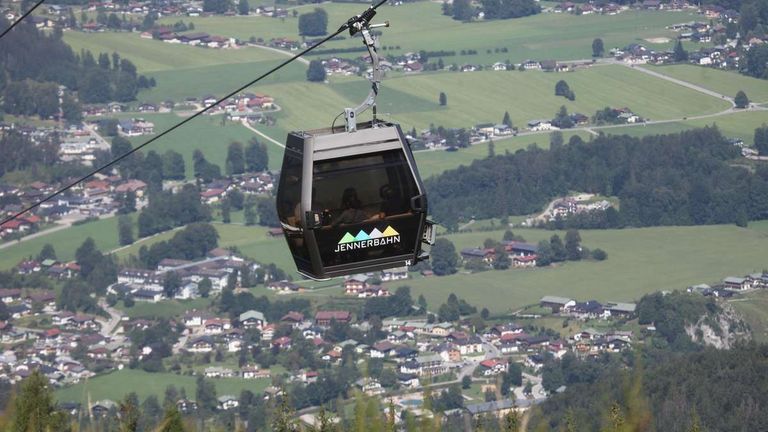 Eine Seilbahn mit der Aufschrift Jennerbahn schwebt über einem Tal mit Blick auf Häuser und grüne Felder.