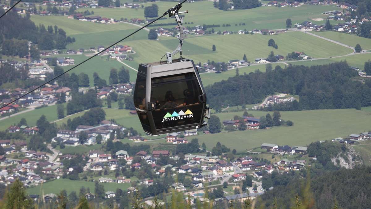Eine Seilbahn mit der Aufschrift Jennerbahn schwebt über einem Tal mit Blick auf Häuser und grüne Felder.